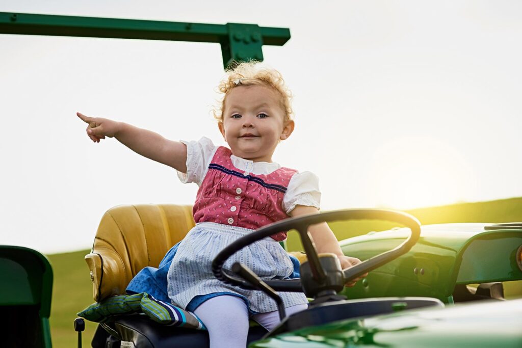 A young child with curly hair, dressed in a traditional dirndl dress, sits on the seat of a green tractor in Montagu and smiles while pointing to the left. The sunset creates a warm glow, perfect for family day trips and memorable activities.
